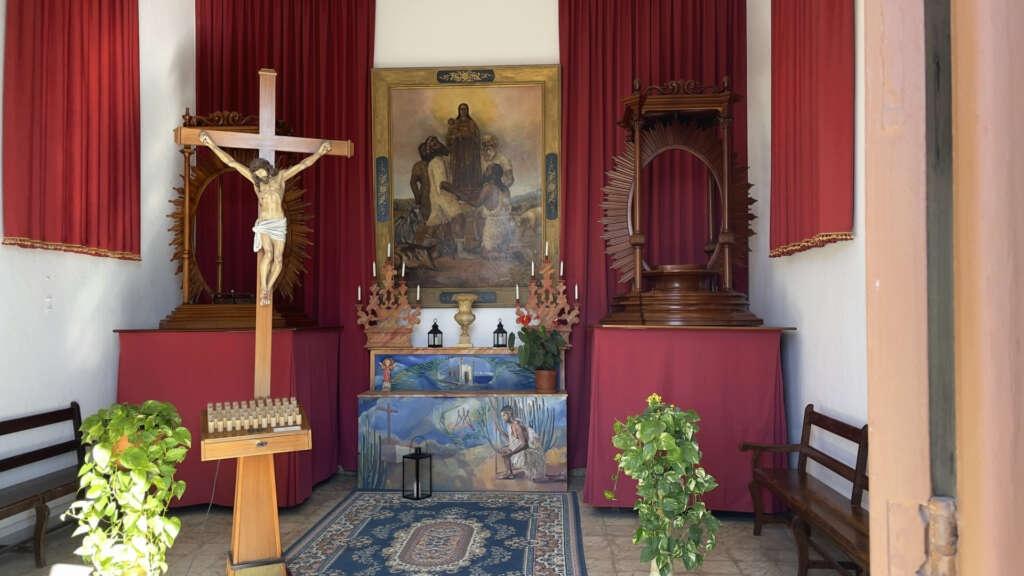 Ein kleiner Innenraum einer Kirche, in dem eine Kreuzfigur aus Holz und mehrere Gemälde mit kirchlichem motiv stehen. Zwei grüne Pflanzen stehen neben einem Teppich.