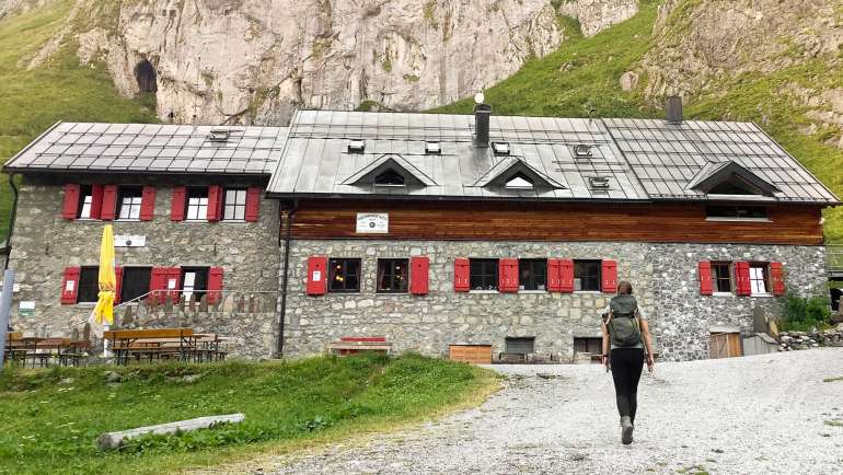 Eine große Berghütte vor Berglandschaft. Eine Wanderin läuft den Weg zur Ravensburger Hütte hoch.