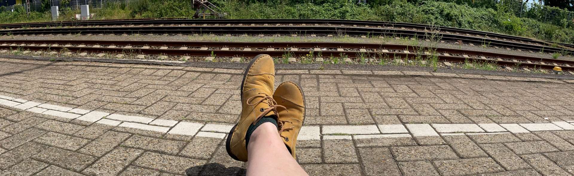 Zwei Füße In Schuhen Auf Einem Bahnsteig, Im Huntergrund Sieht Man Die Bahngleise