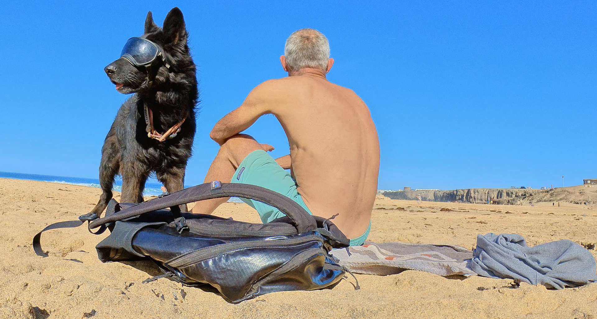 Ein Mann und ein Hund sitzen an einem Sandstrand vor strahlend blauem Himmel. Im Vordergrund sieht man einen geöffneten Rucksack im Sand liegen