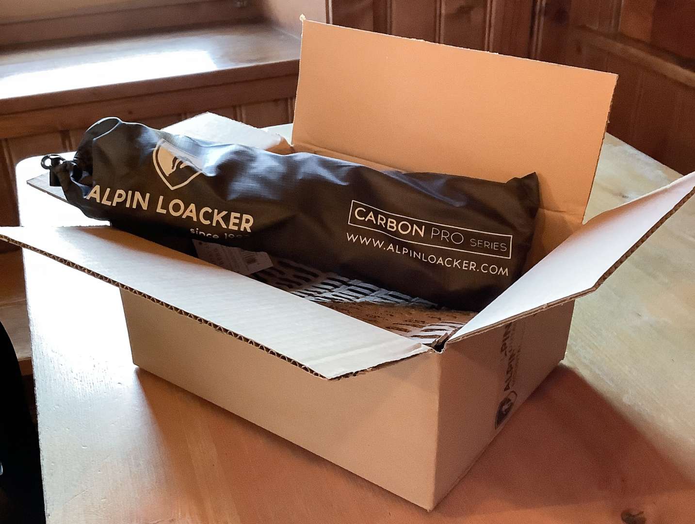 Ein geöffneter Karton auf einem Holztisch, in dem eine schwarze Tasche liegt. Diese trägt in weiß die Aufschrift "Alpin Loacker Caron Pro Series" und die Web-Adresse www.alpinloacker.com