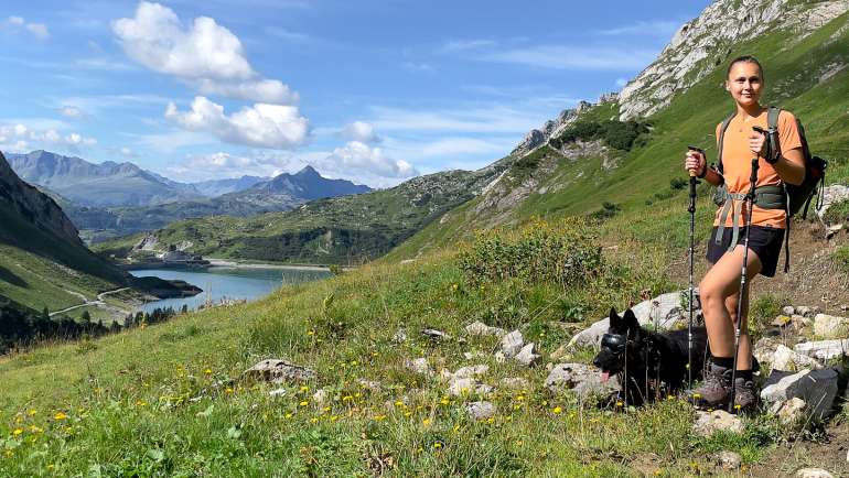 Eine Wanderin und ein Hund stehen vor einer Berglandschaft. Im Hintergrund ist ein Tal mit See zu erkennen.